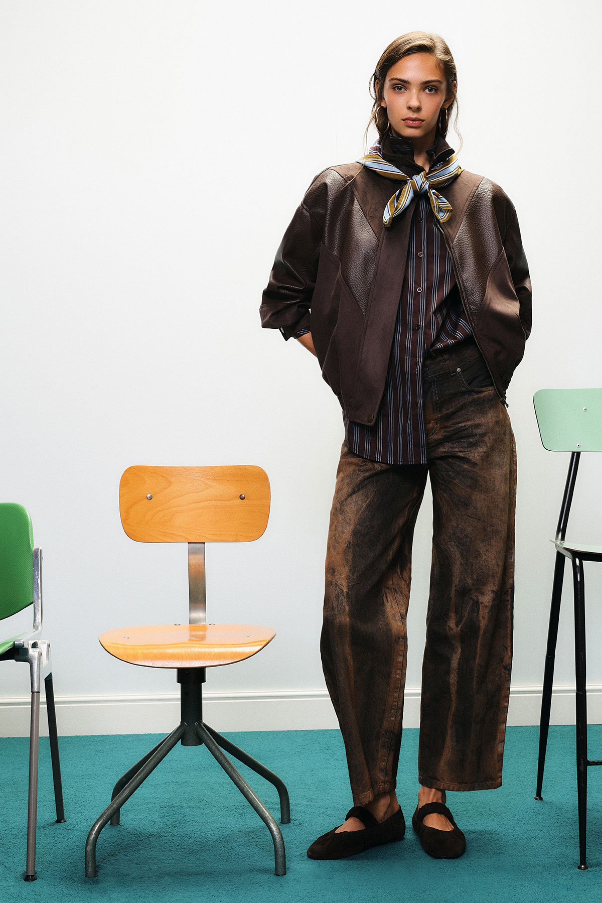 Young woman in brown leather jacket, striped shirt and neck scarf, wide brown trousers and black shoes, standing by chairs