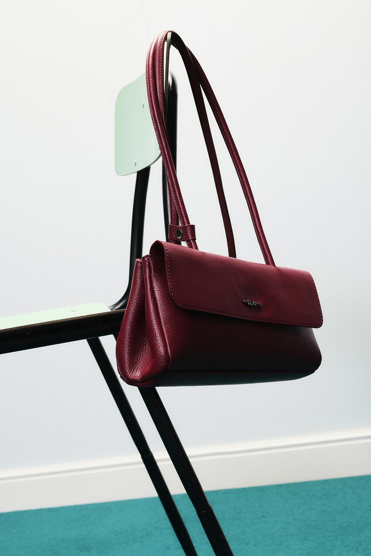 Burgundy leather shoulder bag hanging from a mint-green metal chair in a minimalist room with teal carpet