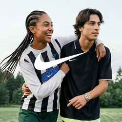 Due giovani camminano su un campo da calcio, uno con il braccio intorno all'altro, entrambi indossano abbigliamento sportivo e sorridono.