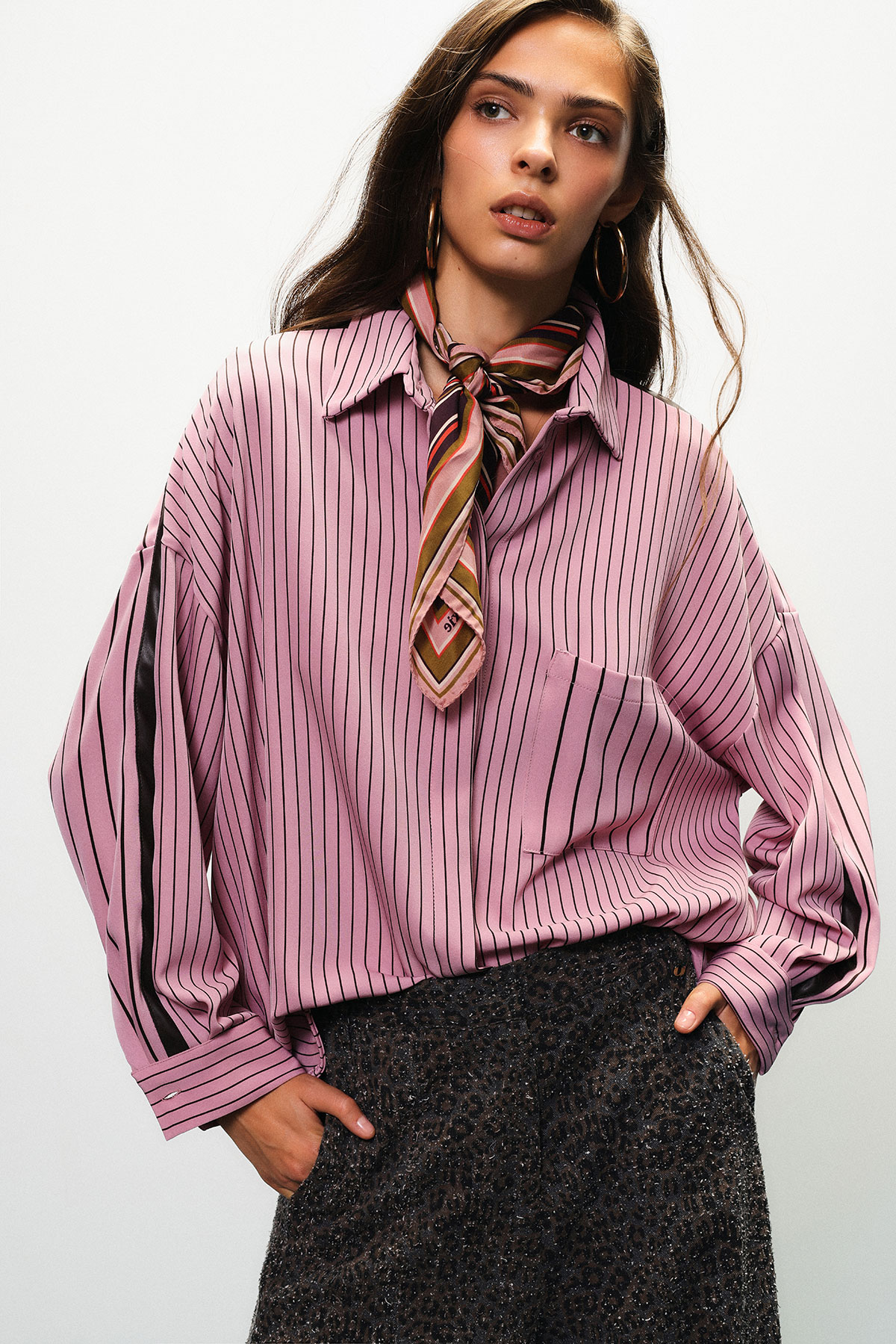 Woman wearing a pink pinstripe oversized blouse with a patterned silk neck scarf and hoop earrings, hands in pockets of a textured dark skirt, studio portrait