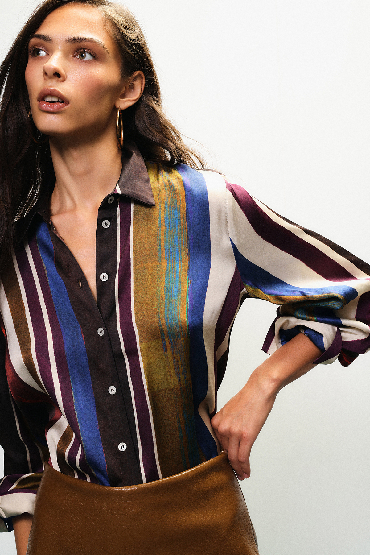 Woman model in multicolour striped silk blouse and brown skirt, hoop earring, hand on hip pose — contemporary women's fashion