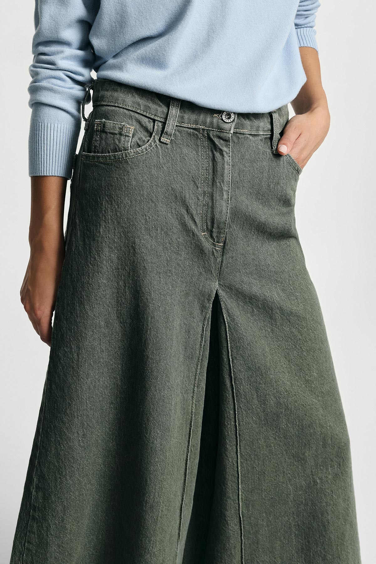 Close-up of person wearing light blue jumper and high-waisted olive-green denim skirt with centre front slit and hand in pocket
