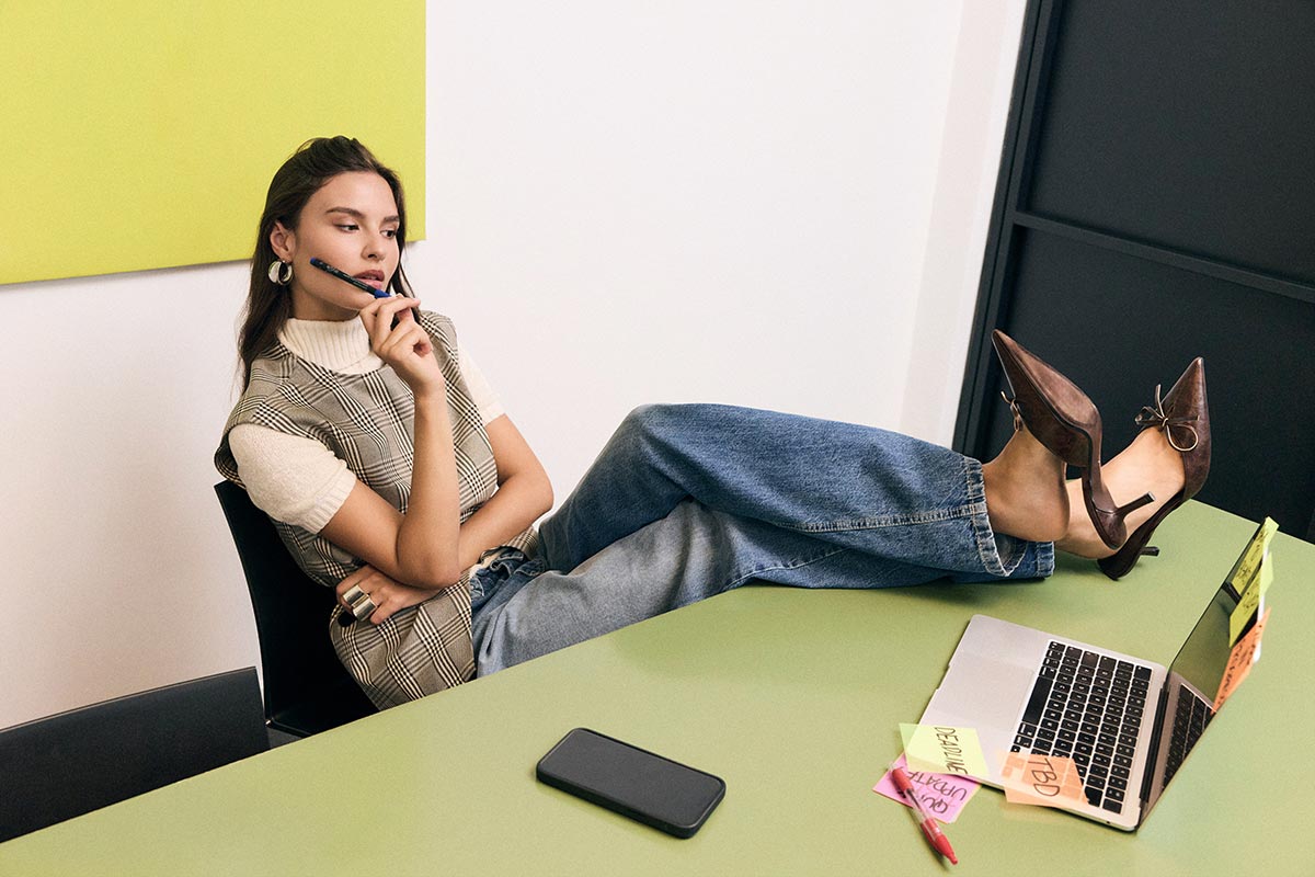 Young woman relaxing with feet on green desk, pen to mouth, laptop with sticky notes and smartphone nearby — casual office work scene