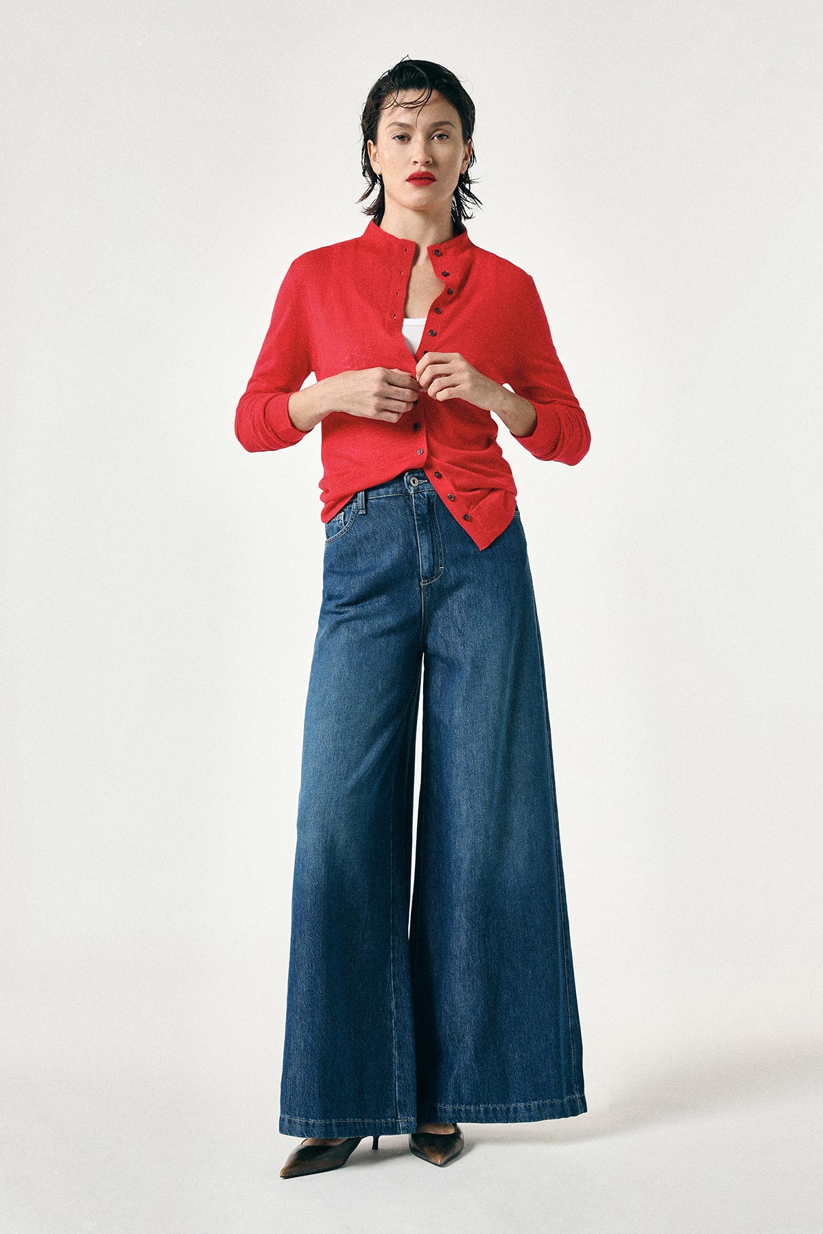 Woman in red buttoned cardigan over white top, high-waisted wide-leg blue jeans and black heels on a plain light background