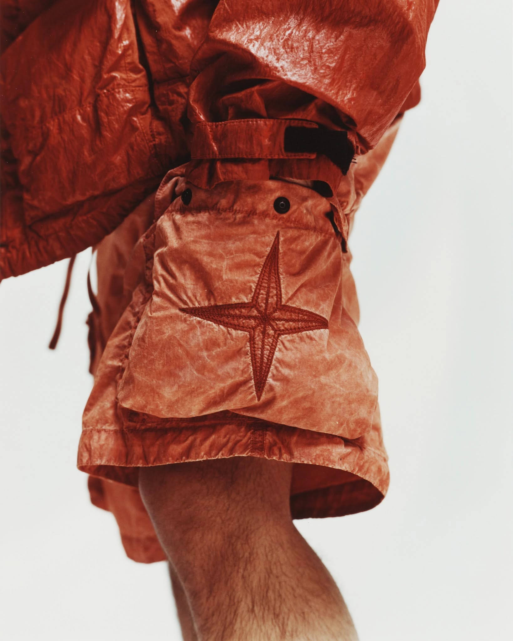 Close-up of rust-coloured crinkled nylon shorts and jacket showing an embroidered star patch above a bare leg