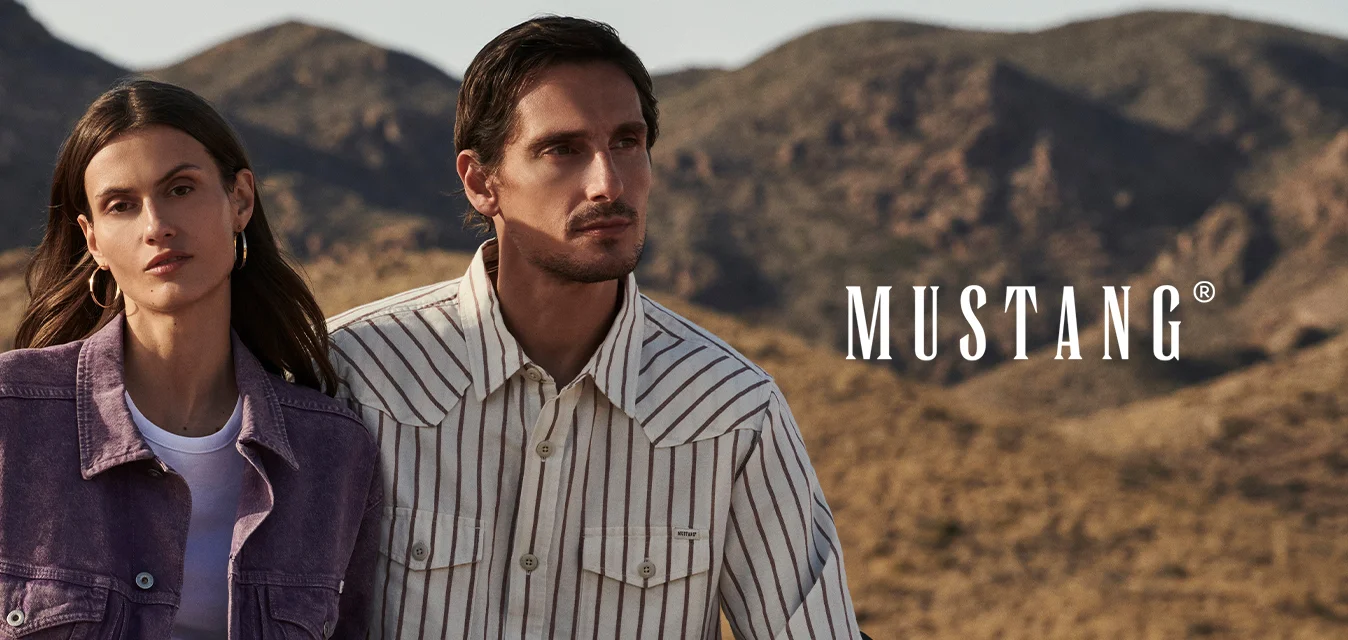 Giovane donna e uomo seduti sul tetto di un veicolo nel deserto, indossano camicie casual a righe viola e bianche con montagne sullo sfondo.