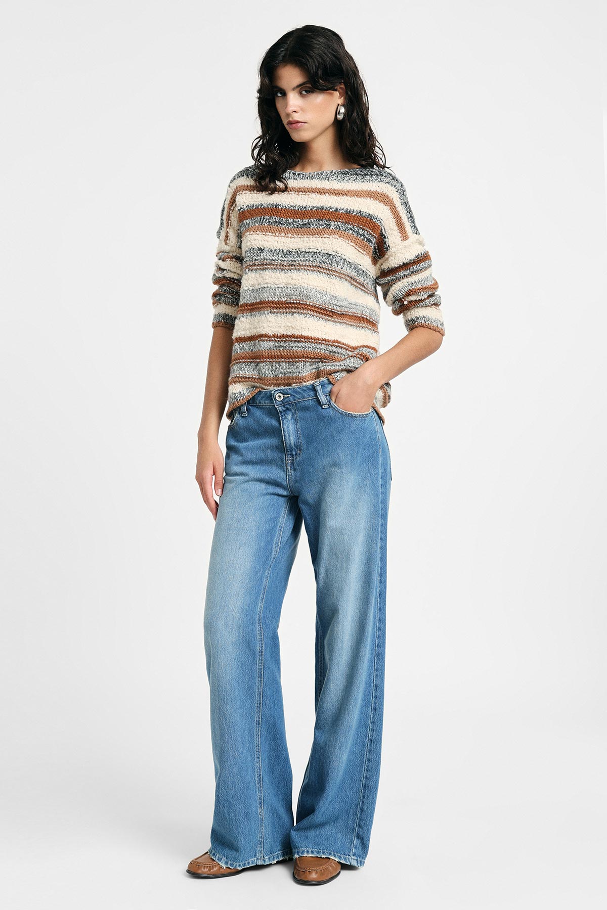 Model in striped beige-brown cosy sweater and wide-leg blue jeans, standing with one hand in pocket on white background