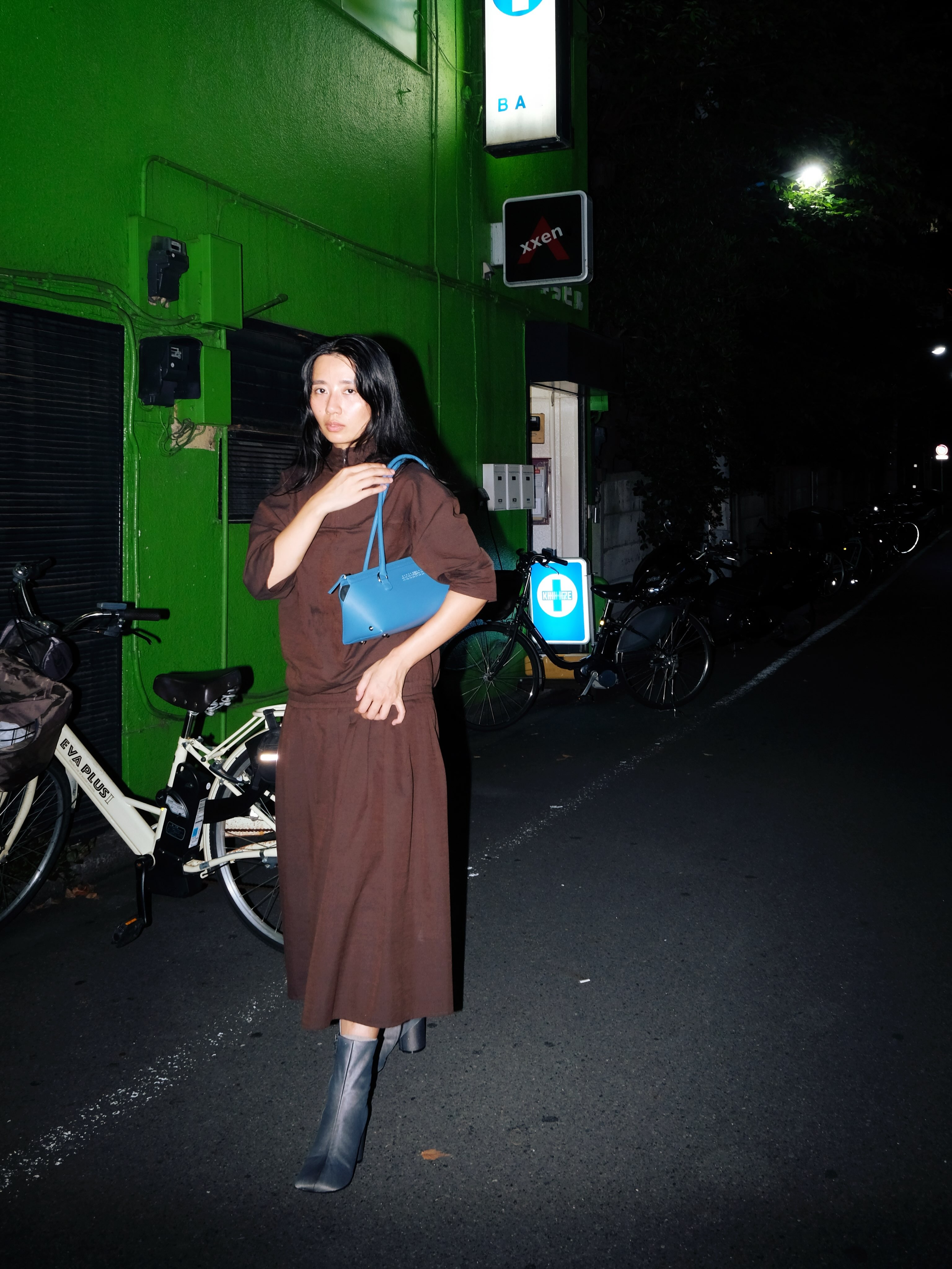Woman in brown dress and grey ankle boots holding small blue handbag, standing on dimly lit street beside green building and parked bicycles