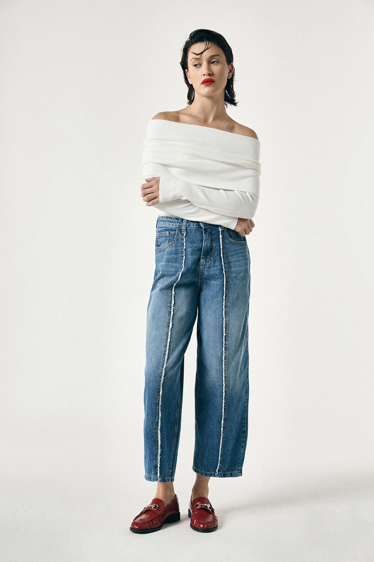 Fashion model in an off-shoulder white sweater, wide-leg frayed-seam blue jeans and red loafers on a plain background