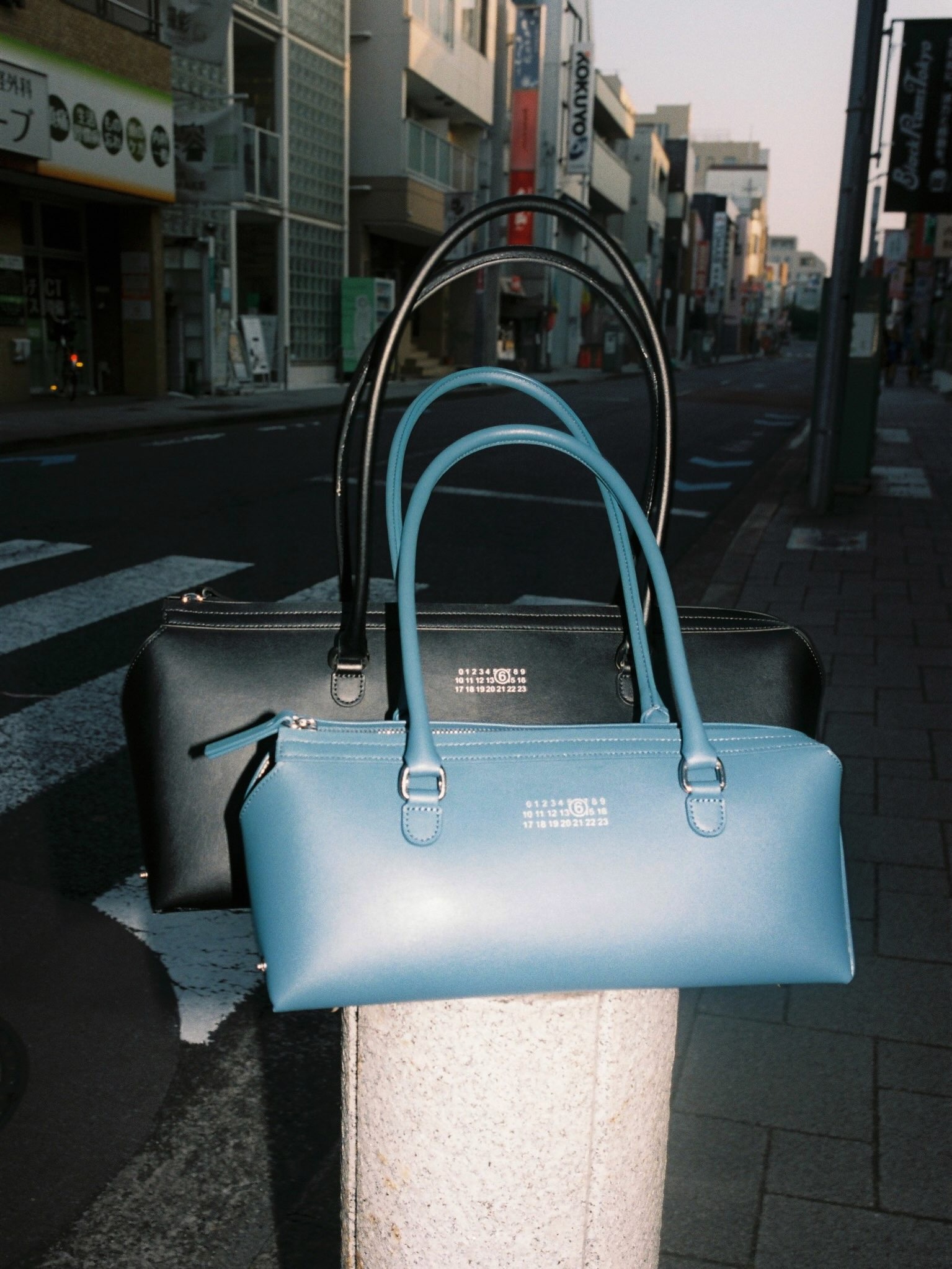 Two rectangular leather handbags, blue and black, displayed on a pavement bollard on a quiet city street
