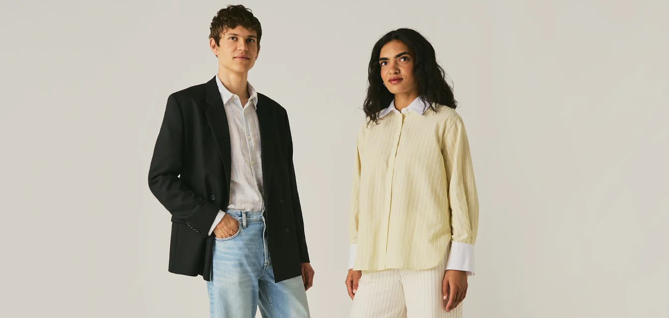 Due persone sono in piedi su uno sfondo chiaro uniforme; una indossa un blazer nero con jeans, l'altra una camicetta beige con pantaloni bianchi e sandali.