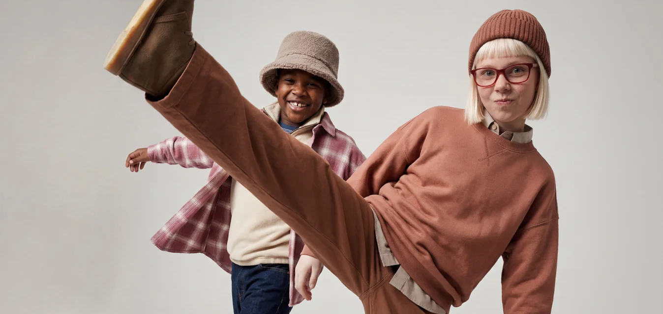 Due bambini che ballano in abiti casual, uno che dà un calcio alto indossando un completo marrone e un berretto, l'altro che sorride con una camicia a quadri e un cappello.