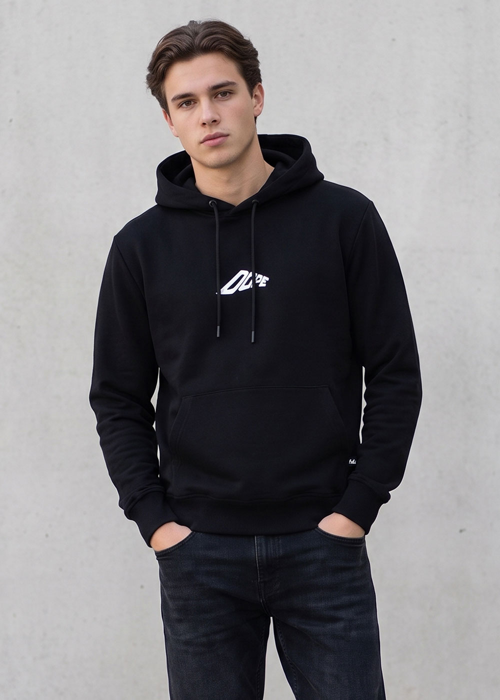 Young man modelling black hoodie with small white logo, hands in pockets against plain grey wall