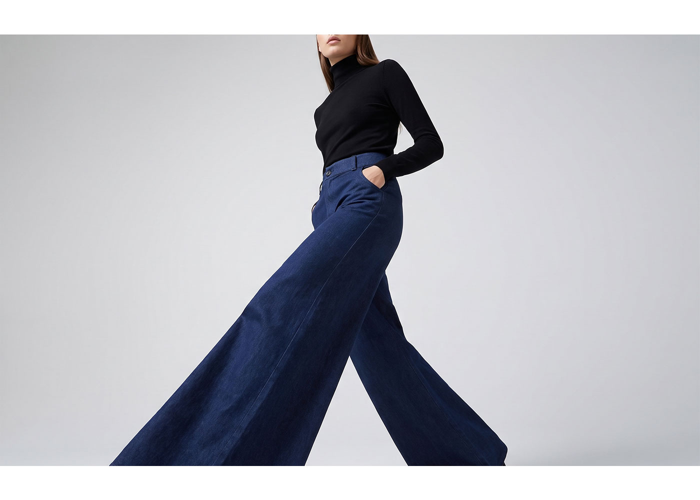 Model wearing high-waisted navy wide-leg trousers and black turtleneck jumper, walking with hands in pockets against a plain pale background