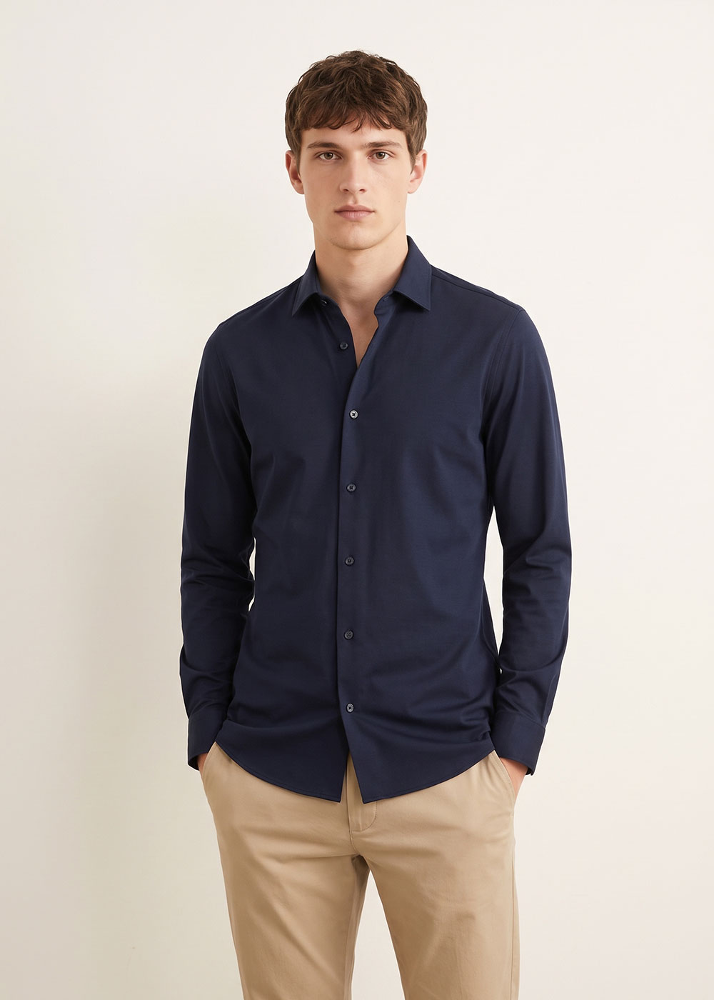 Young man wearing a navy slim-fit long-sleeve shirt and beige chinos, standing against a plain light background