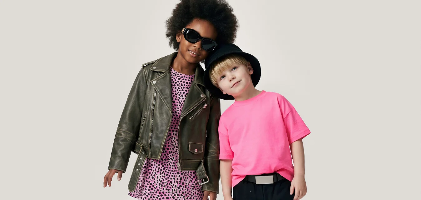 Due bambini stanno vicini, uno indossa occhiali da sole, una giacca di pelle e un vestito; l’altro indossa una camicia rosa acceso e un cappello nero.