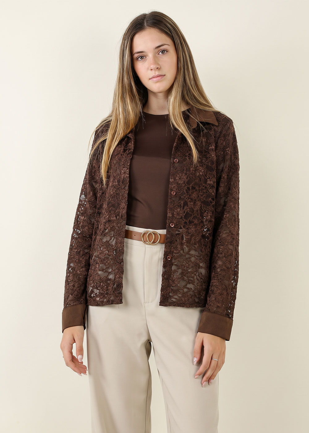 Woman wearing a brown lace shirt over a brown top and beige trousers with a double-ring belt on a pale background