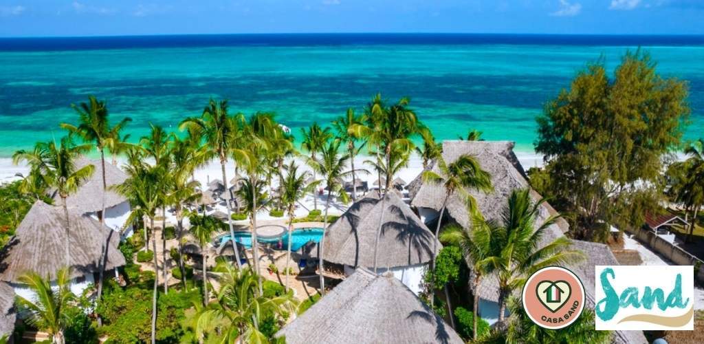 Aerial view of a tropical beachfront resort with thatched-roof bungalows, palm trees, pool and turquoise sea; Casa Sand logo