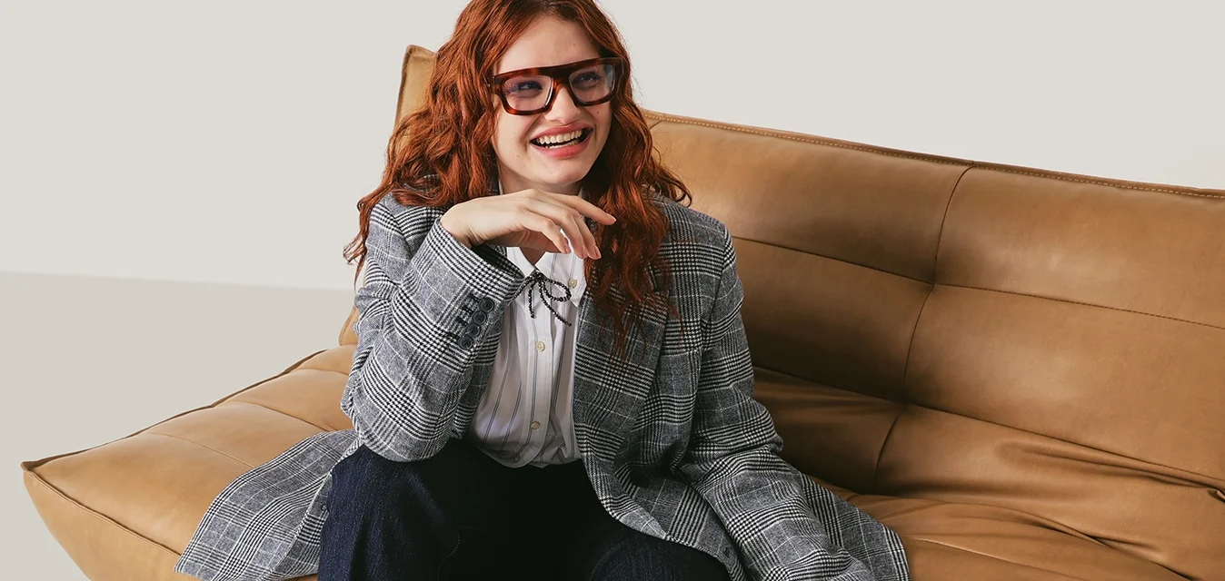 Donna dai capelli rossi, con occhiali, blazer a quadri e pantaloni scuri, seduta su una moderna poltrona in pelle color cuoio, con una borsa sulla sinistra, sorridente.