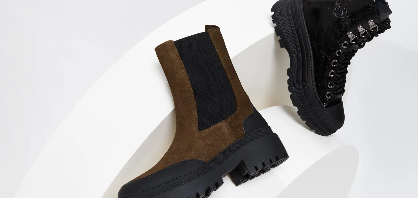 Stivale alla caviglia in suede marrone con pannelli elastici neri, suola nera spessa; stivale nero con lacci di superficie lucida e occhielli metallici.