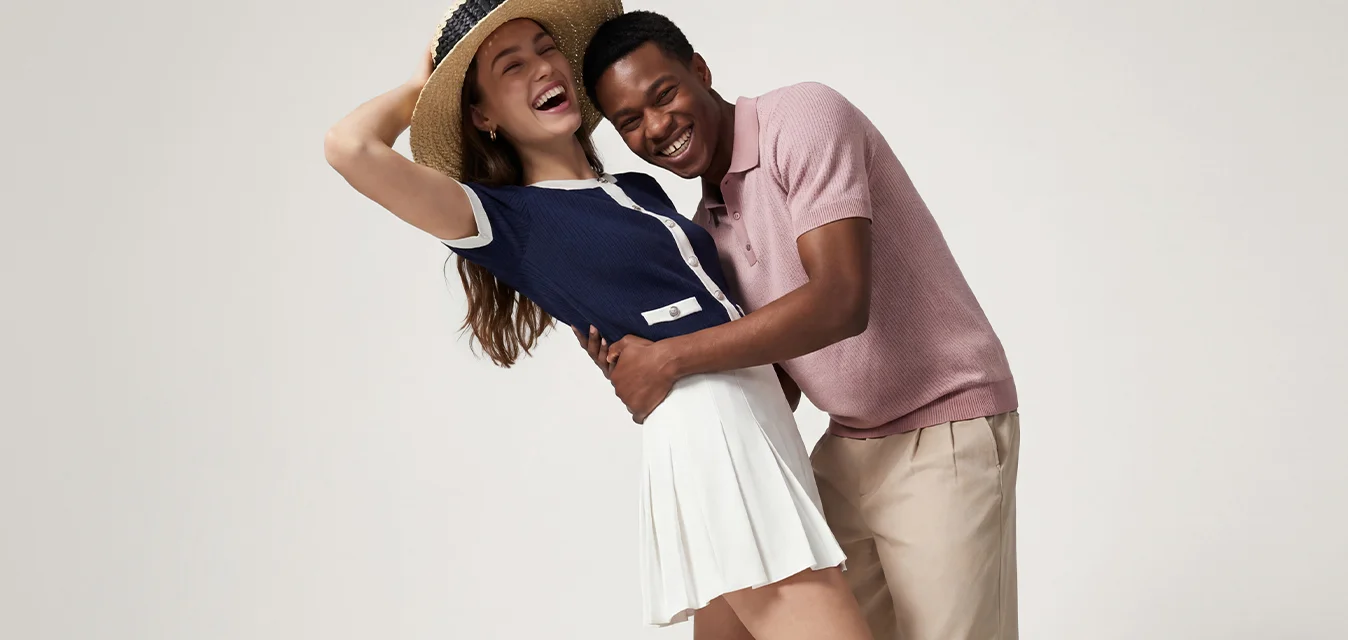 Giovane uomo e donna che ridono e si abbracciano, donna con cappello, top blu navy e gonna bianca, uomo con camicia rosa e pantaloni beige su sfondo bianco.