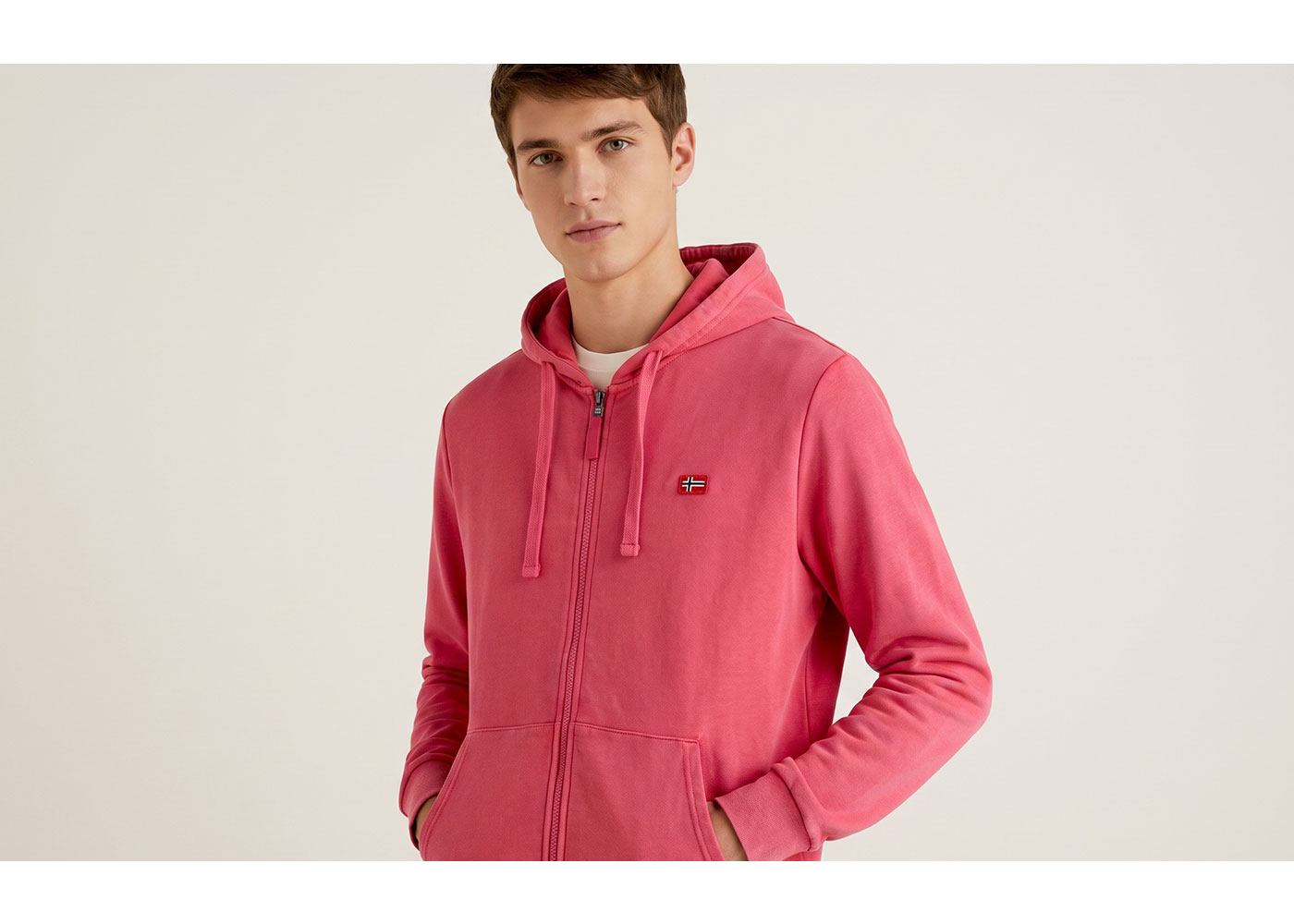 Young man wearing pink zip-up hoodie with small flag patch, hands in pockets, neutral background