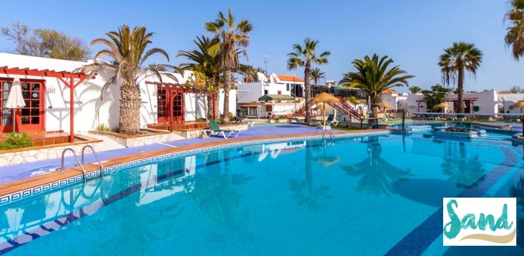Outdoor resort swimming pool with clear blue water, sun loungers and palm trees beside white Mediterranean-style bungalows
