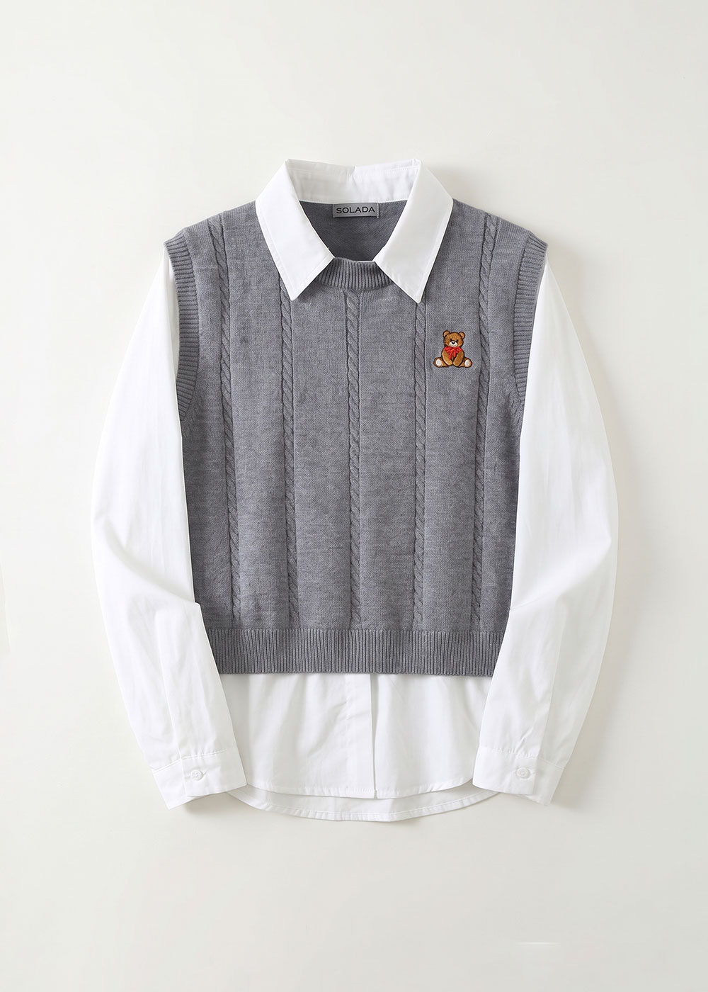 Grey cable‑knit sleeveless sweater vest layered over white collared shirt, small embroidered teddy bear on left chest