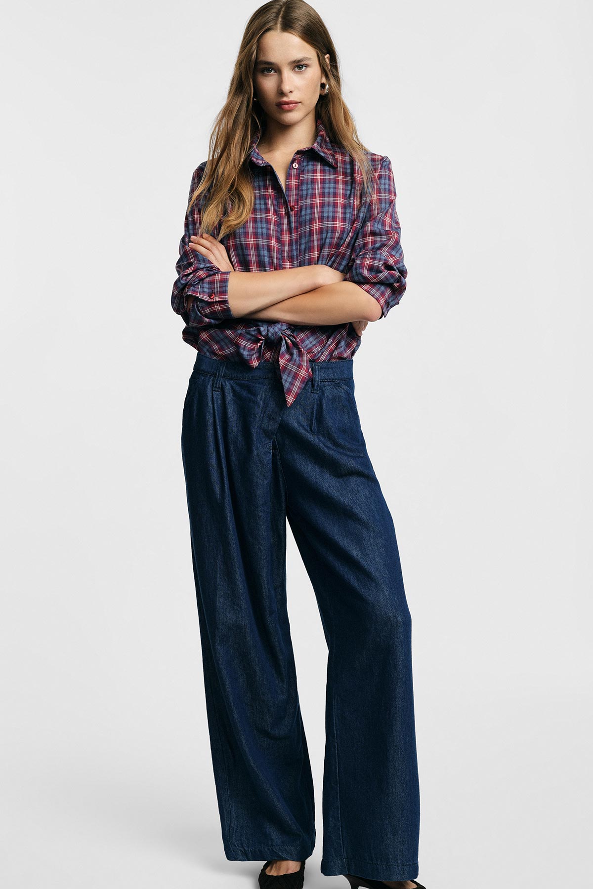 Woman wearing tied red plaid shirt and high-waisted wide-leg dark denim trousers, arms crossed against plain background