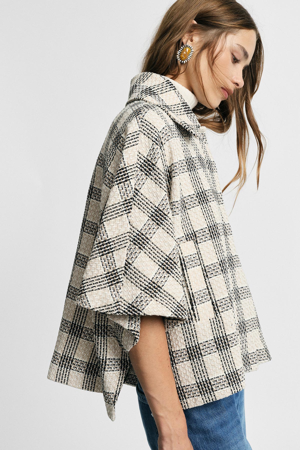 Young woman modelling a cream and black checked cropped cape coat with blue jeans, side profile highlighting a large statement earring
