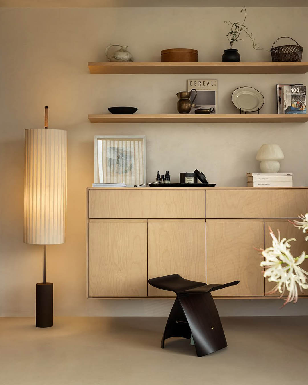 Minimalist living room with floating light-wood sideboard, shelves, tall pleated floor lamp and dark wooden stool