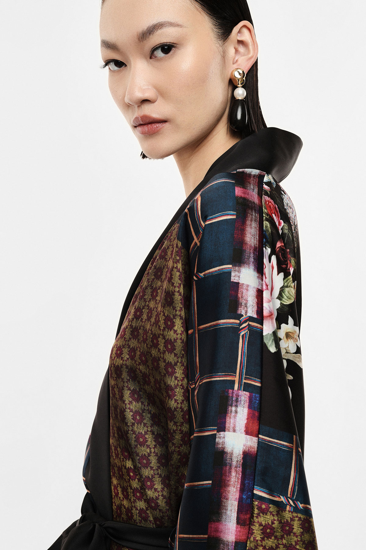 Close-up of woman wearing a patterned kimono-style jacket with floral and checked prints and a large pearl drop earring against a plain background