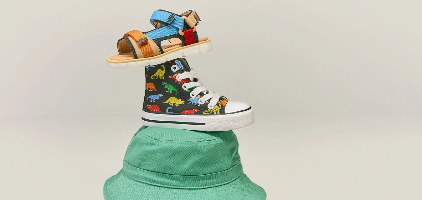 Accessori impilati per bambini: cappello a secchiello verde, sneaker alta nera con dinosauri colorati e sandalo blu e arancione con chiusura a strappo sopra.