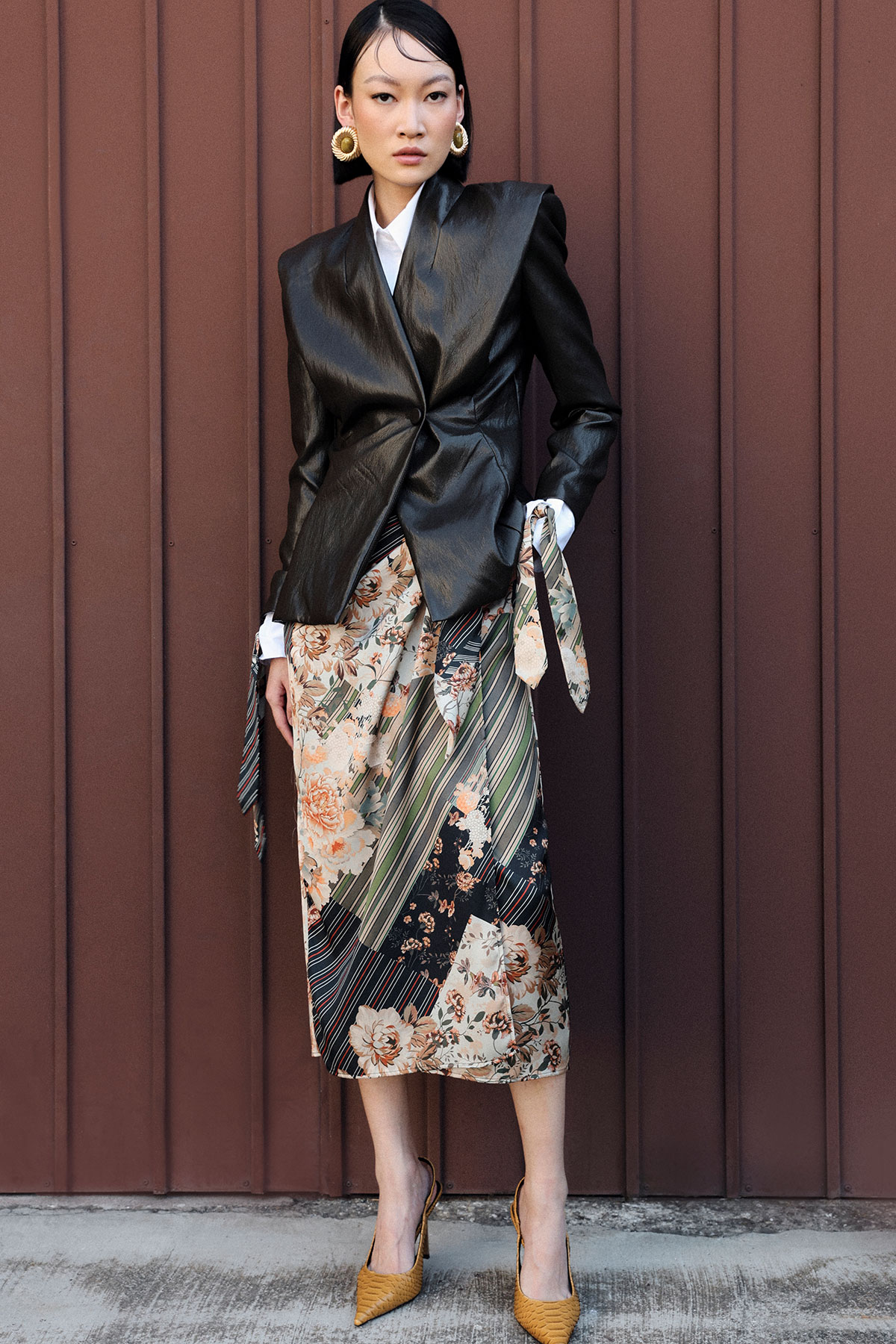 Woman in structured black leather blazer, floral midi skirt, mustard heels and gold hoop earrings against brown metal wall