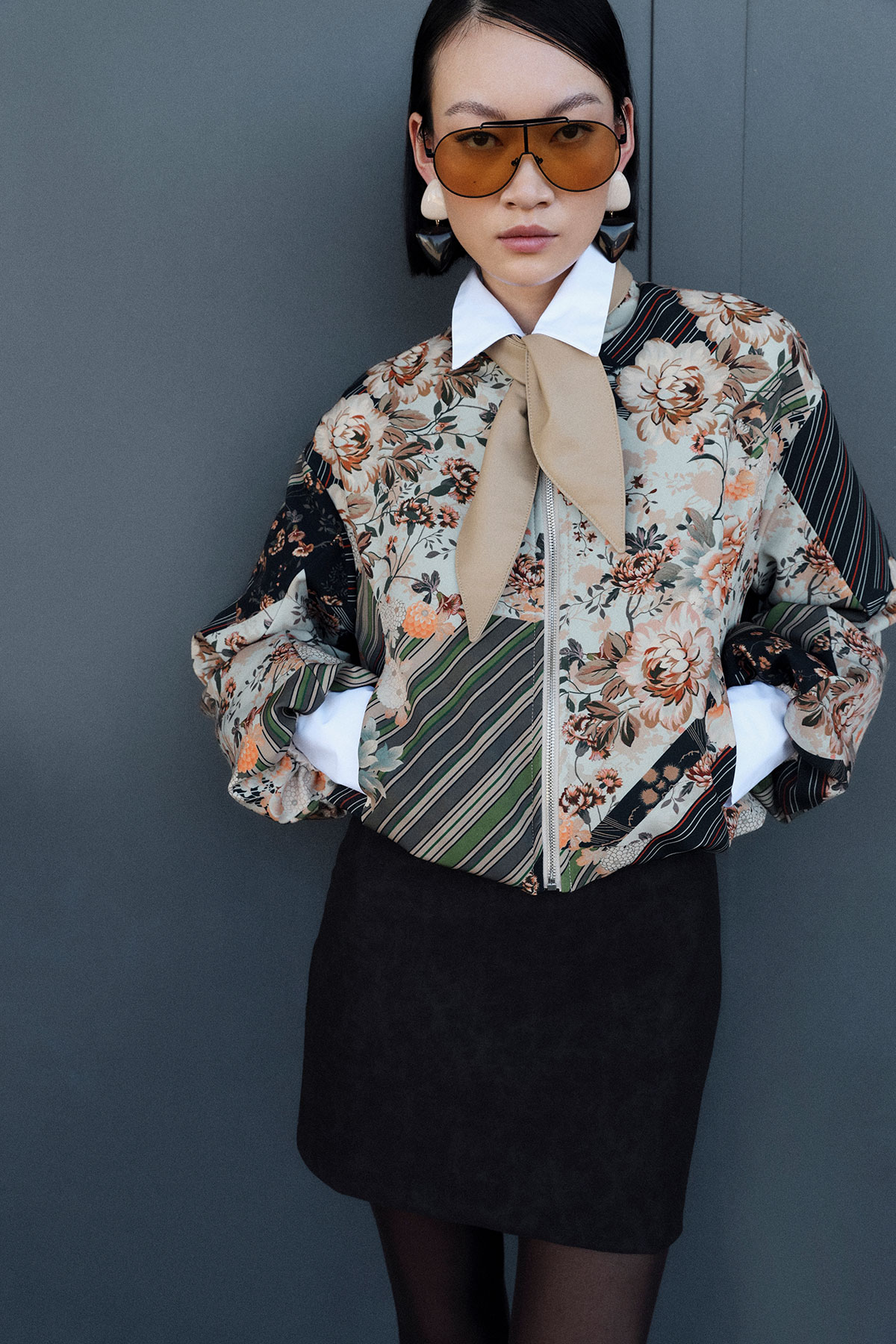 Woman wearing floral and striped bomber jacket with white collar and beige bow, oversized sunglasses, statement earrings, black skirt