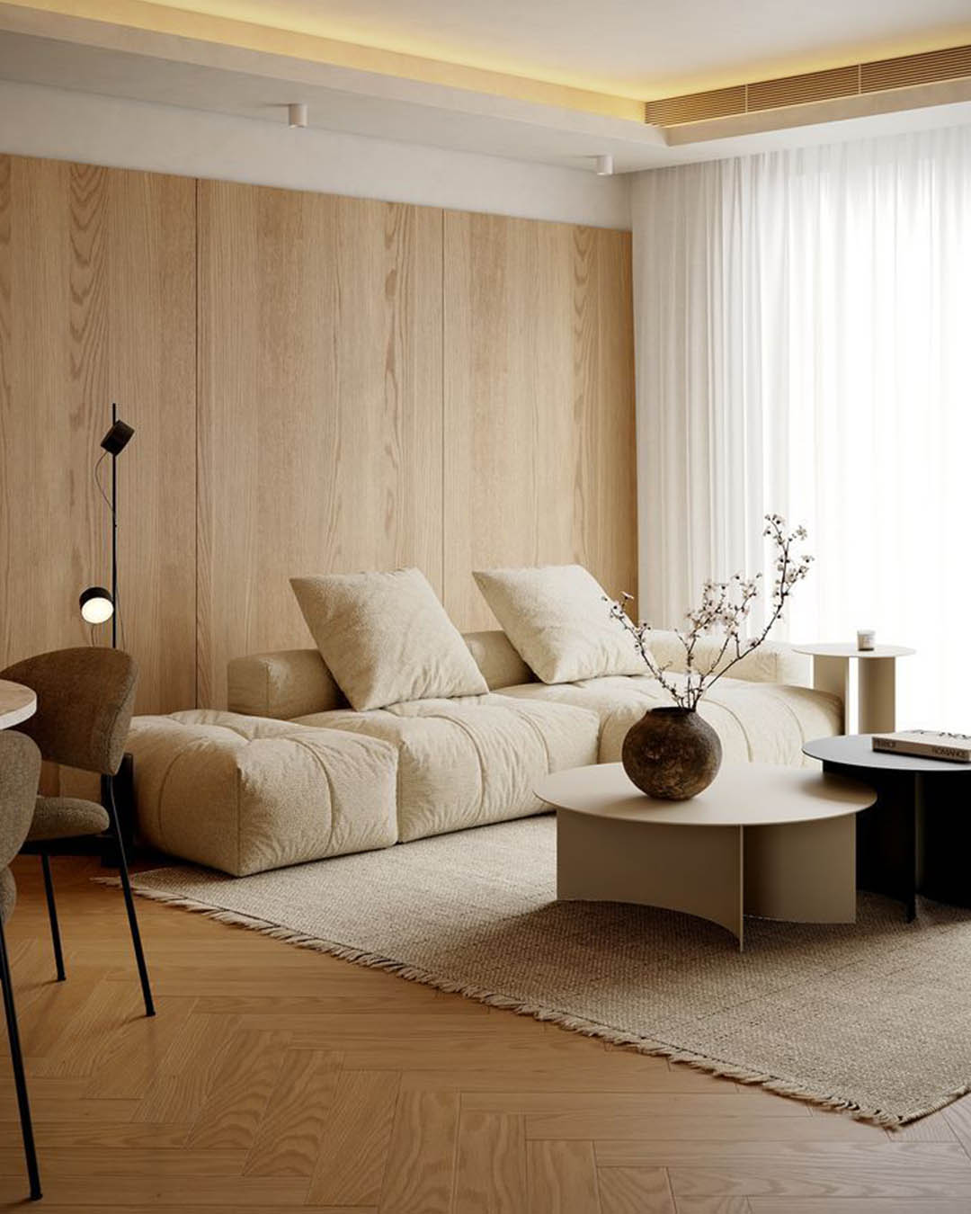 Minimalist neutral living room with low beige modular sofa, wood‑panelled wall, round coffee tables, clay vase with branches and sheer curtains