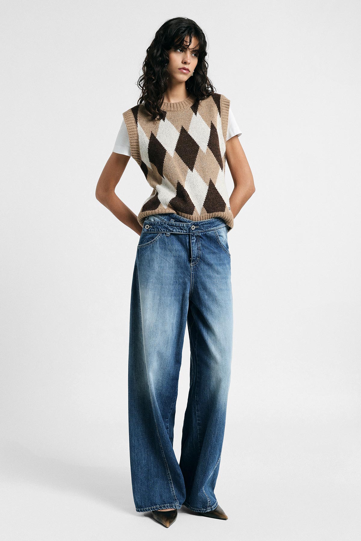 Woman standing in studio wearing beige and brown argyle sweater vest over white T‑shirt with wide‑leg blue jeans and pointed shoes
