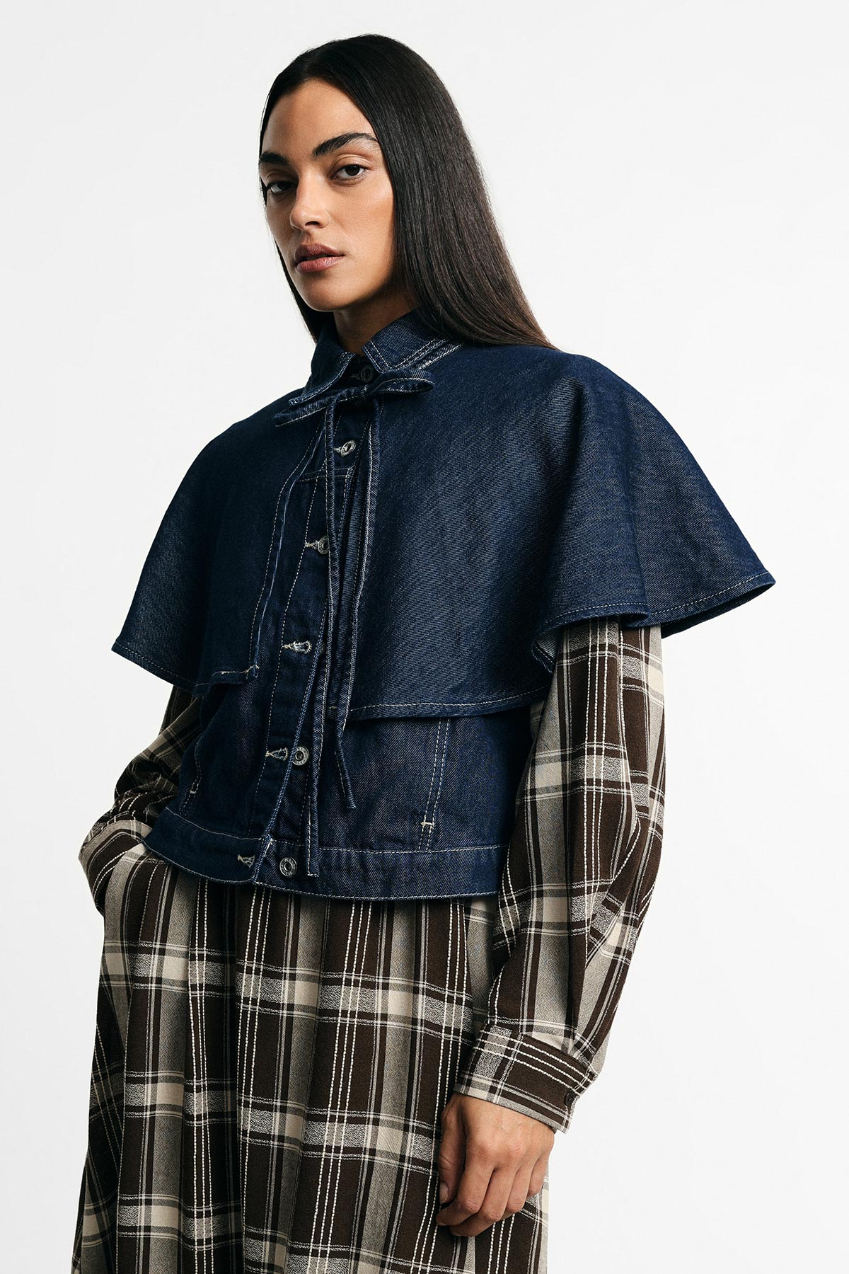 Woman wearing navy denim capelet over brown plaid dress, hand in pocket, standing against a plain white background