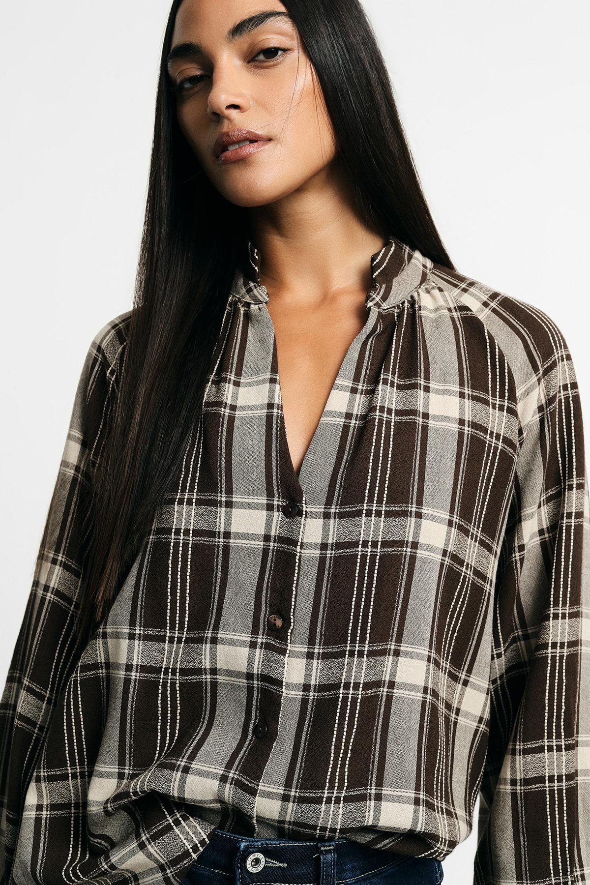 Close-up of a woman with long straight hair wearing a brown and cream checked blouse tucked into dark jeans on a white background