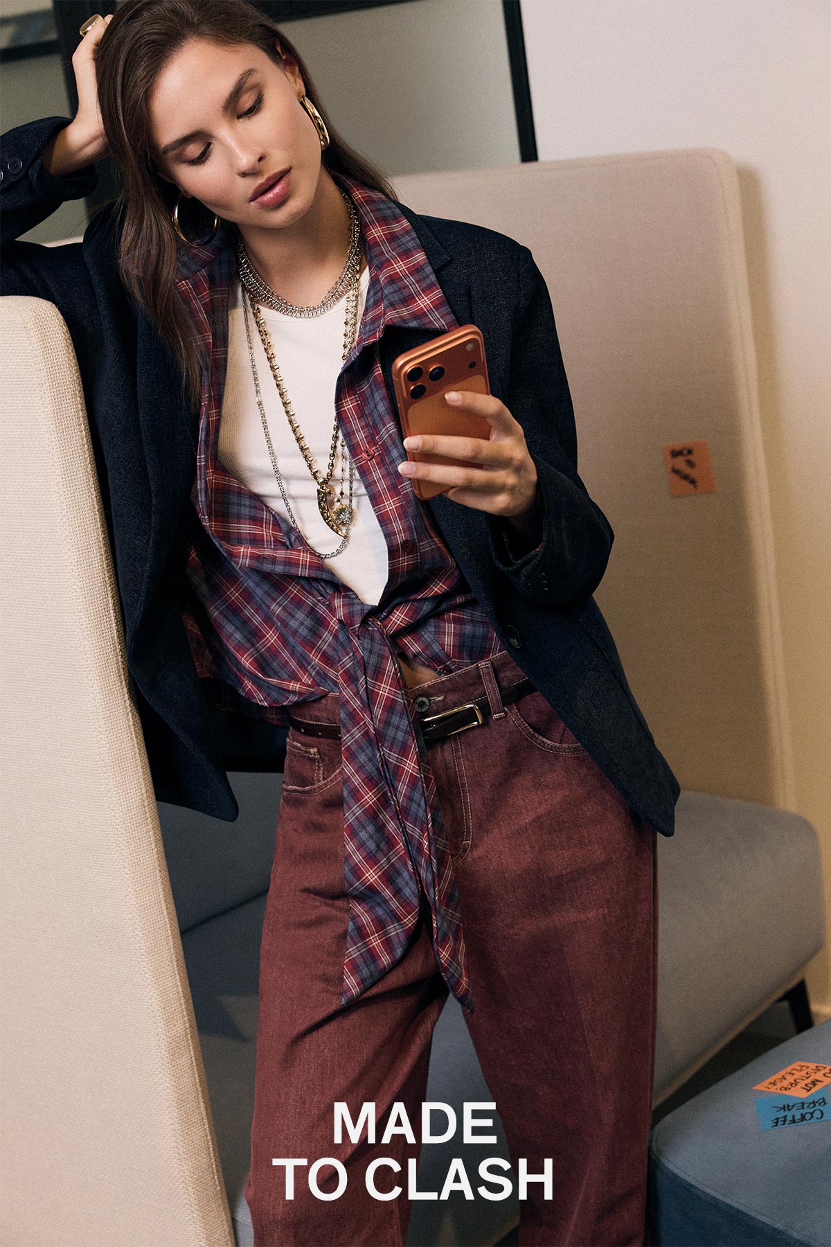 Woman in layered plaid shirt, blazer and jewellery checking a smartphone while leaning on a beige armchair, caption 'MADE TO CLASH'
