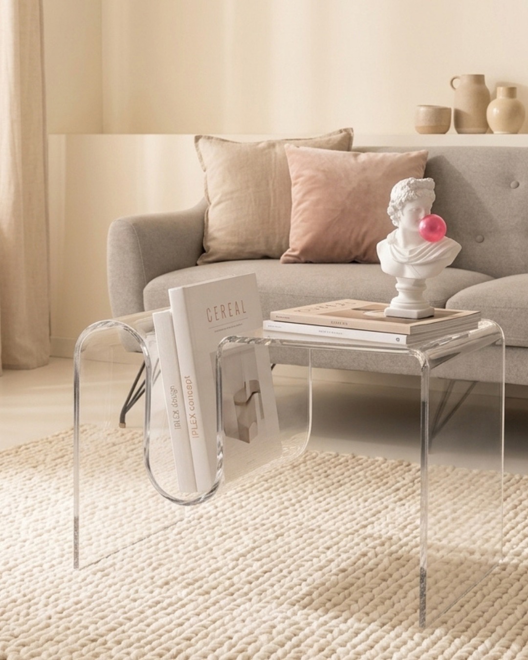 Acrylic curved coffee table with built-in magazine rack holding design books, sculpted bust blowing pink bubblegum, neutral sofa and textured rug in sunlit living room