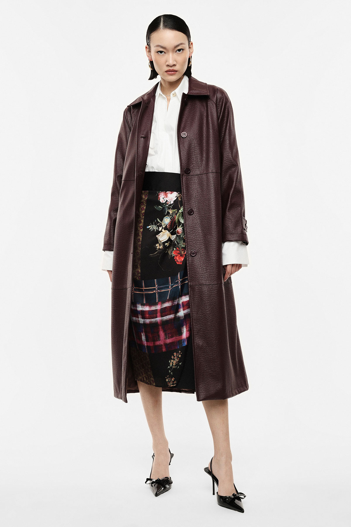 Model wearing long burgundy leather coat over white blouse and floral-plaid midi skirt with black heels