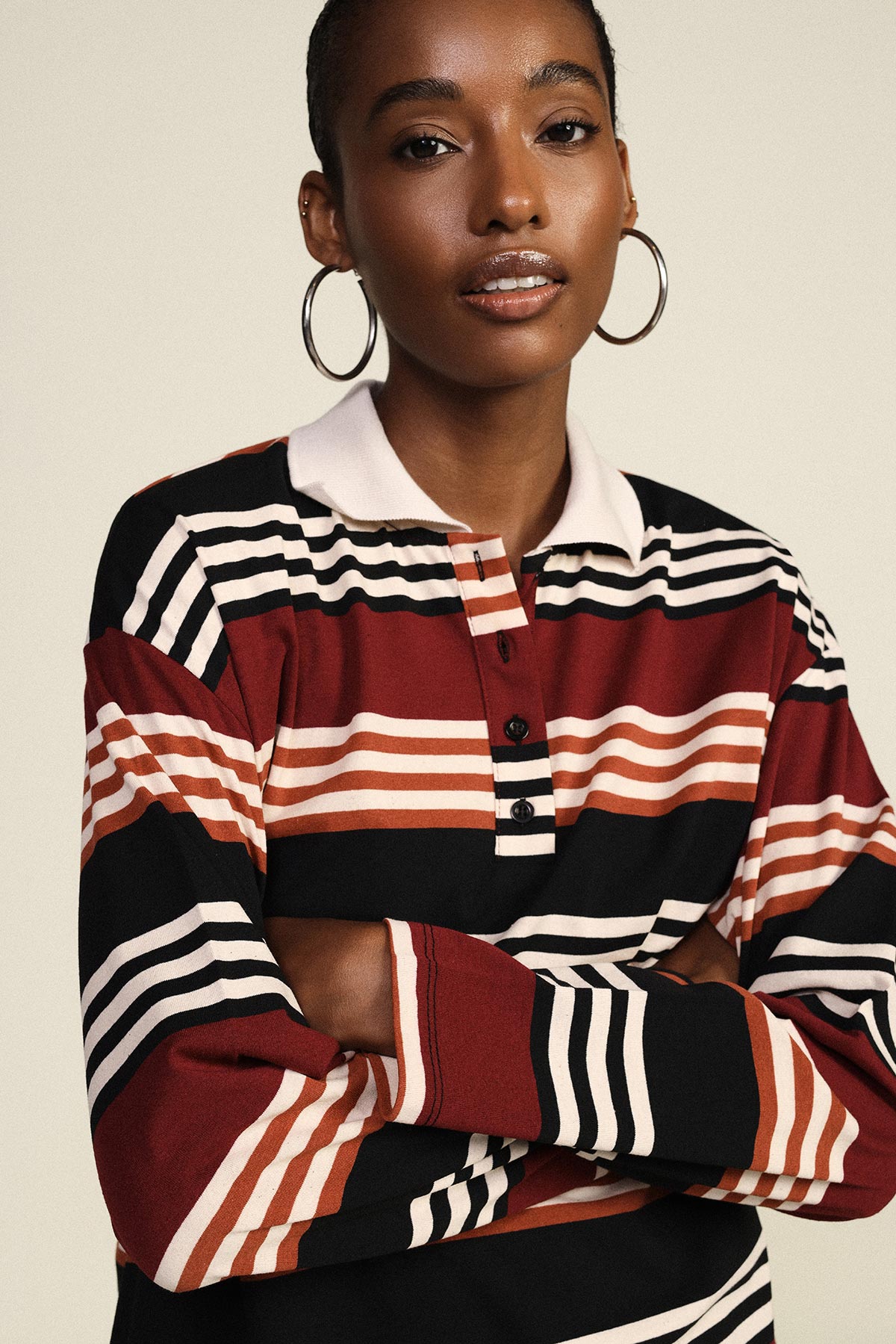 Woman modelling maroon, black and white striped rugby shirt with hoop earrings and arms crossed against a neutral background