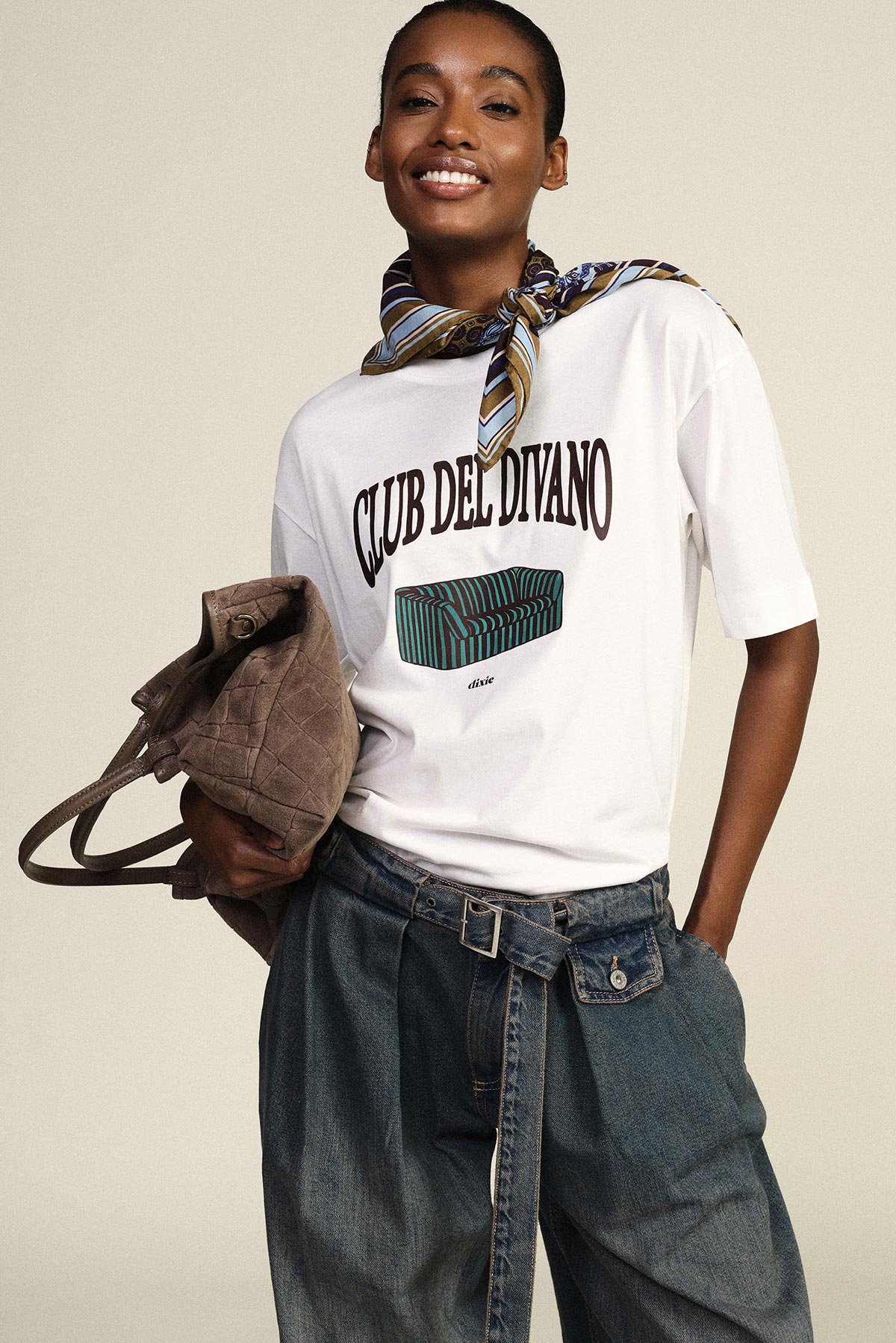 Smiling model wearing white 'CLUB DEL DIVANO' graphic T-shirt, striped neck scarf, high‑waist denim trousers with belt, holding suede bag