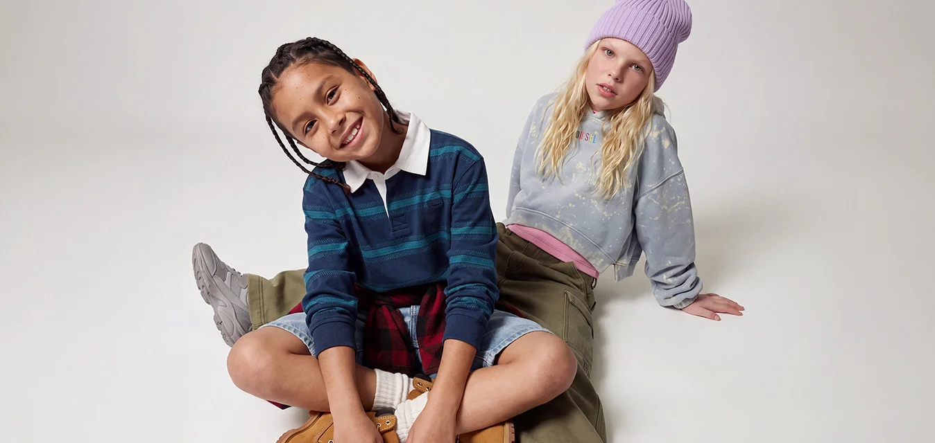 Due bambini seduti sul pavimento, uno sorride con un maglione a righe e stivali, l’altro con un berretto e abiti casual appoggiato indietro.