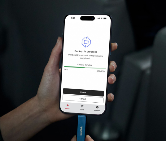 Primo piano delle mani che collegano un'unità telefonica SanDisk Maritime Blue con USB-C a un telefono, con il telefono che mostra una schermata «Backup in Progress» al 50% di completamento.