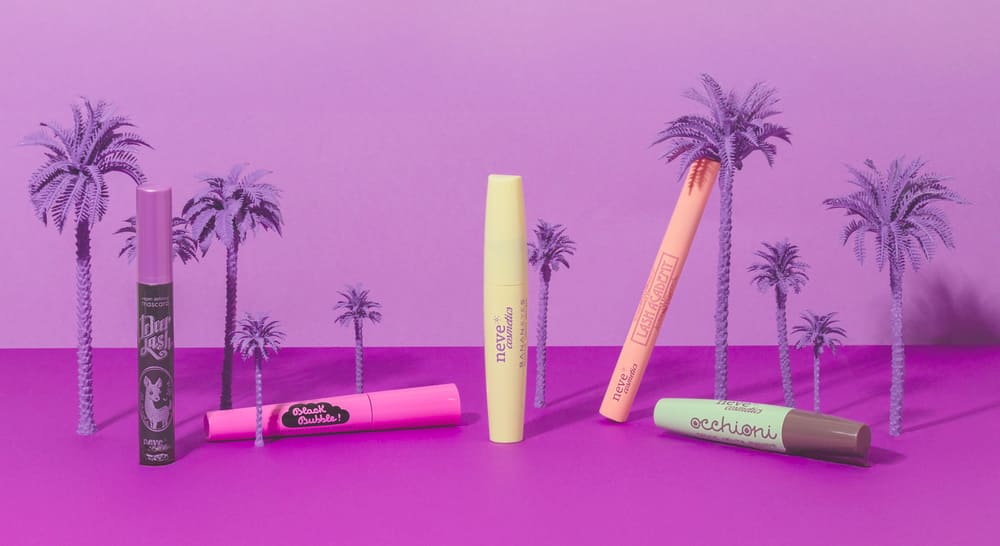 Assortment of colourful mascara tubes on a vivid purple surface with miniature tropical palm trees, beauty cosmetics display