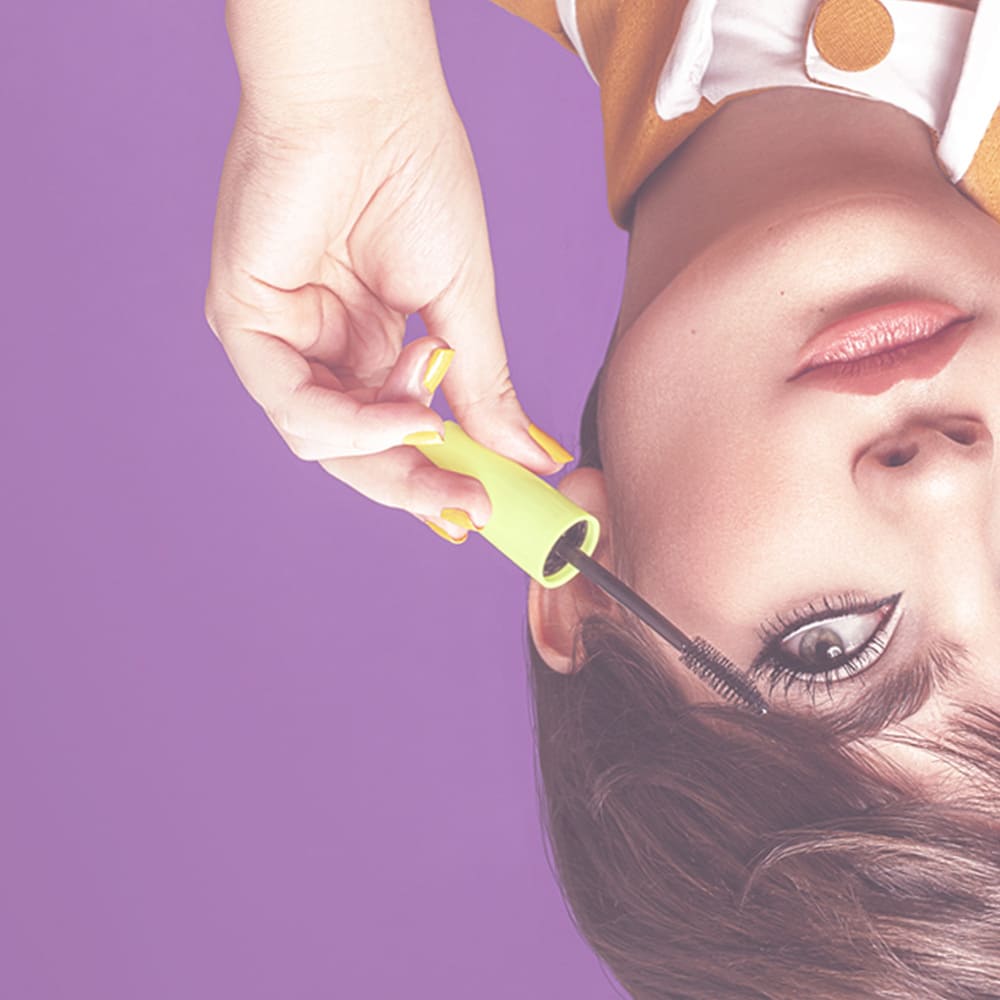 Upside-down close-up of a woman applying mascara to her upper lashes with a green wand against a purple background
