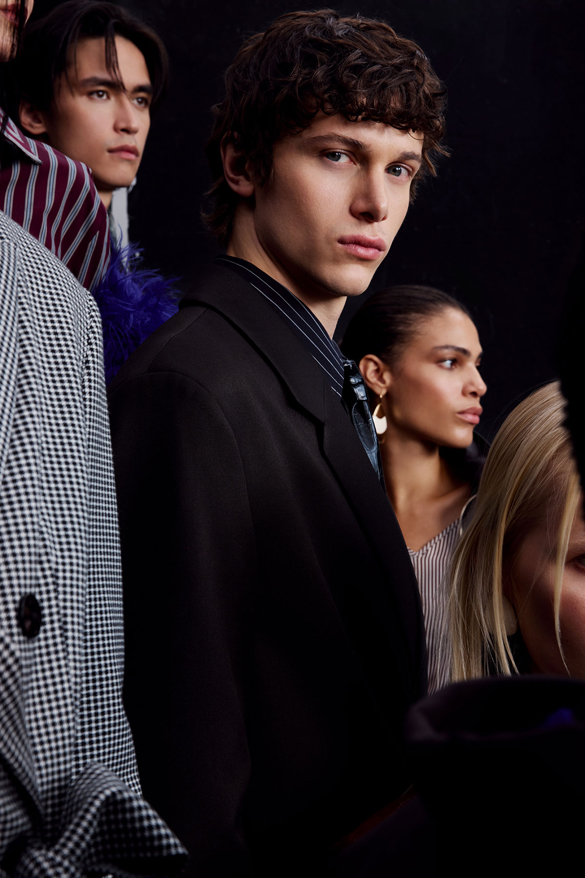 Group portrait of diverse young fashion models in tailored coats, central male model gazing at camera against a dark backdrop