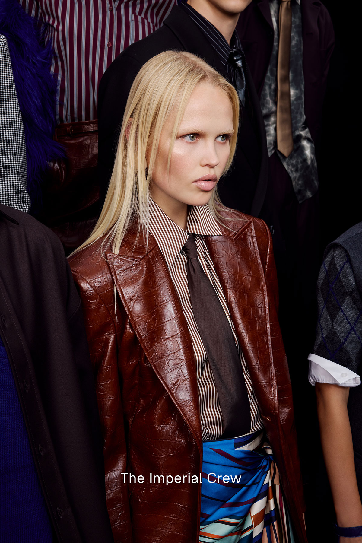 Young blonde model in a brown leather blazer, striped shirt and dark tie standing among a stylish group